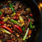 Homemade Kung Pao Beef with tender beef strips, red chilies, scallions, bell peppers, and peanuts in a glossy spicy-sweet sauce, served in a cast iron skillet.