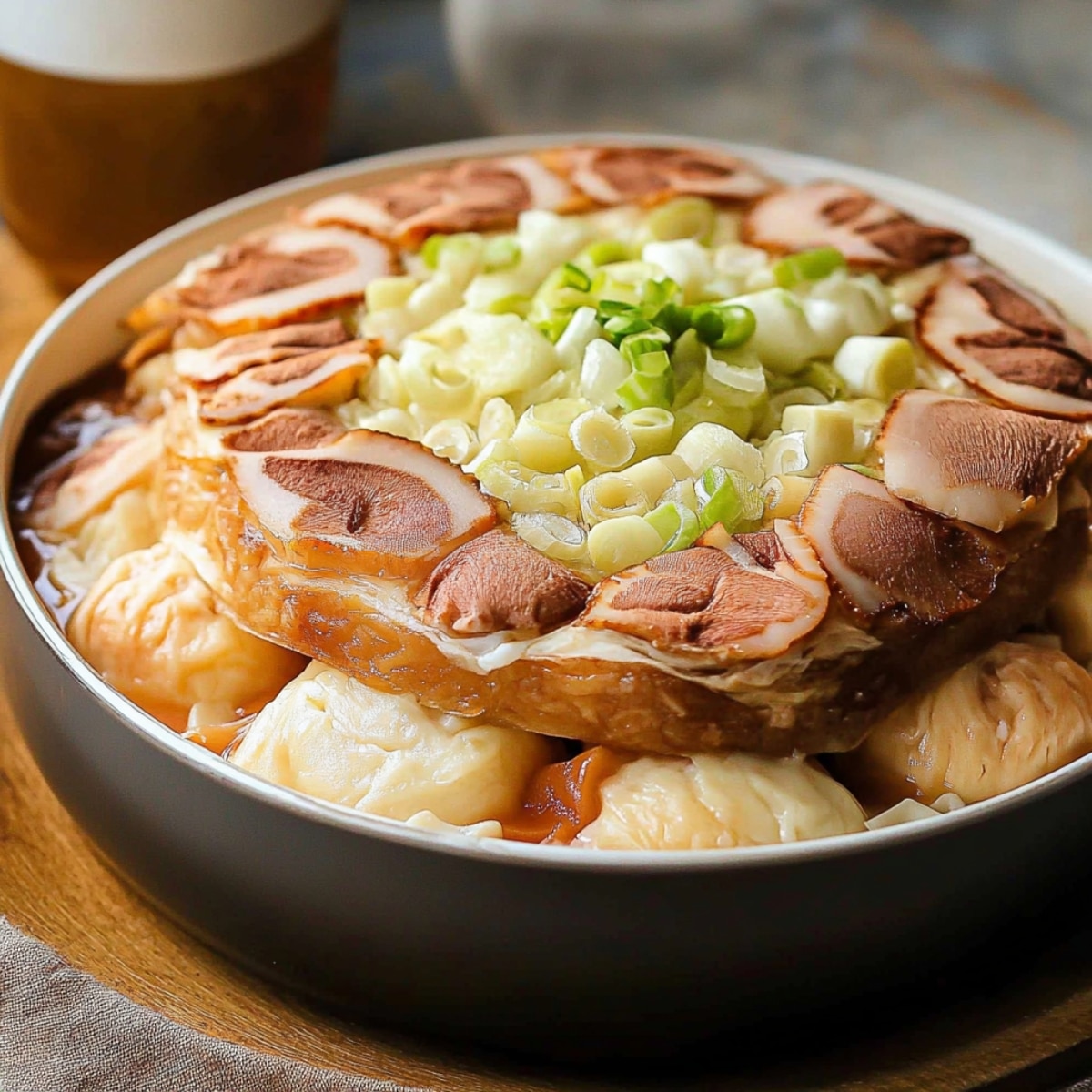 A close-up of a homemade Mille Feuille Nabe in a black pot, with layers of napa cabbage and slices of char siu (roast pork) on top. Fresh green onions garnish the dish, and a golden, clear broth surrounds the ingredients. The soft lighting gives the dish a warm, homemade look.