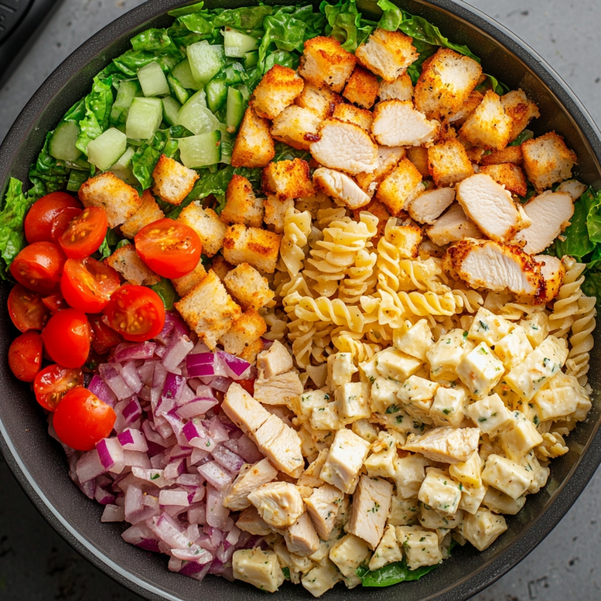 A close-up of a homemade chicken Caesar pasta salad with romaine lettuce, cucumber cubes, cherry tomatoes, red onion, crispy croutons, grilled chicken breast slices, rotini pasta, and cubed chicken coated in creamy dressing, all neatly arranged in a bowl.