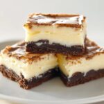 Three homemade vanilla cheesecake brownies stacked on a white plate, with a fudgy chocolate base, creamy vanilla layer, and a golden, slightly cracked top crust, lit softly to highlight the rich, real textures.