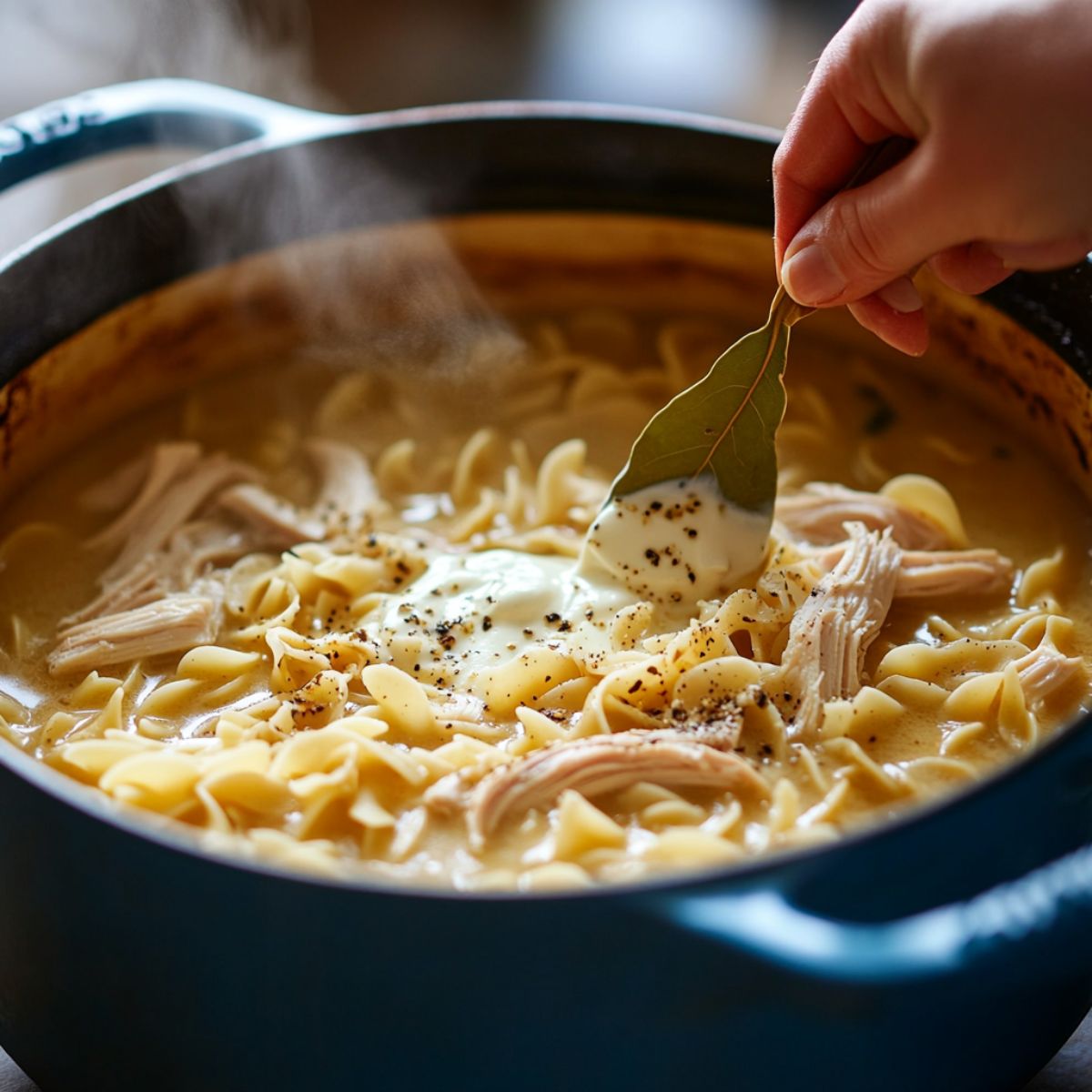 Creamy chicken noodle soup simmering in a blue pot with noodles, shredded chicken, cream, black pepper, and a bay leaf being added by hand.