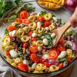 Tortellini pasta salad with tomatoes, mozzarella, olives, spinach, and red onion, tossed with dressing in a glass bowl.