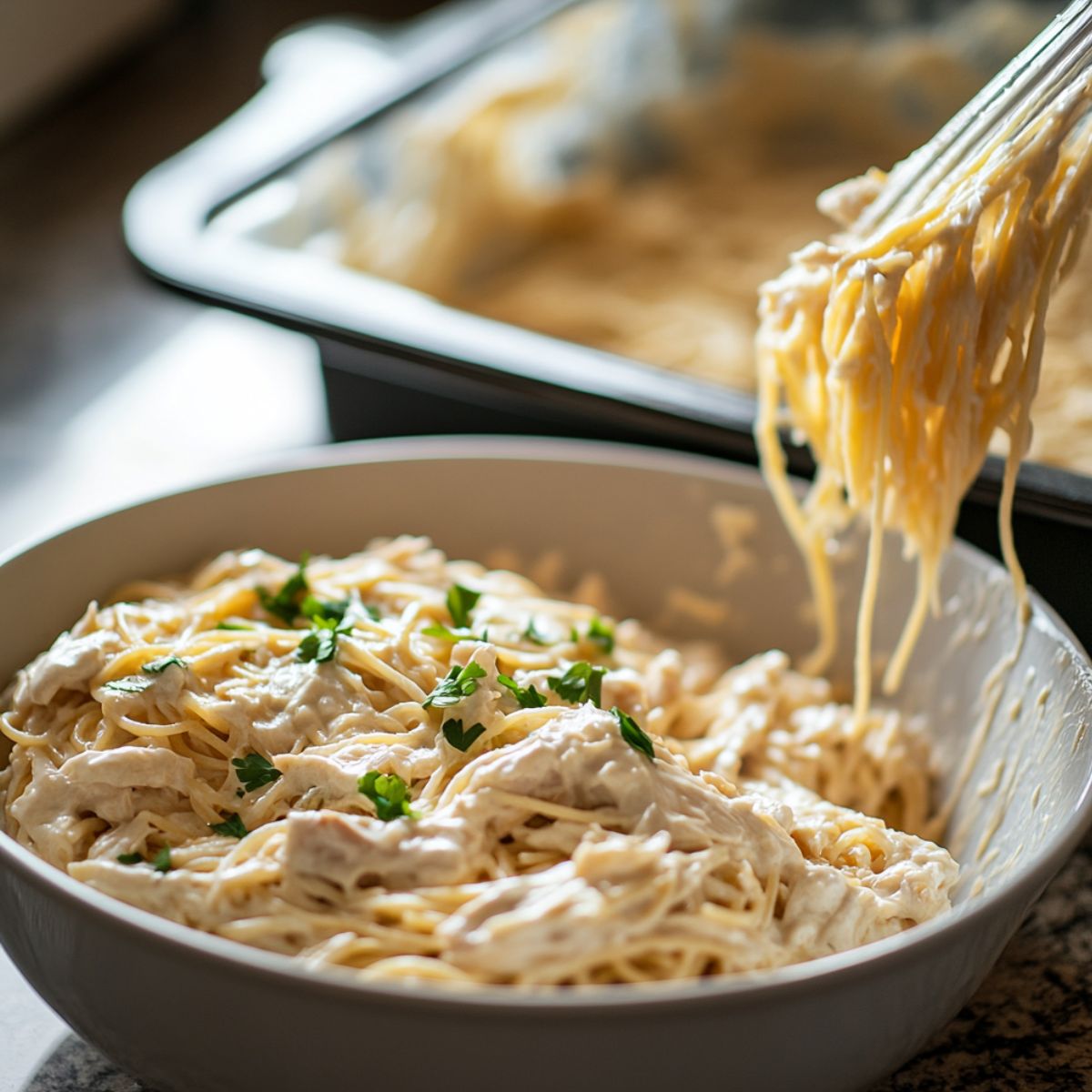 Creamy chicken spaghetti in a bowl topped with parsley, with tongs lifting noodles and a baking dish in the background on a kitchen counter.