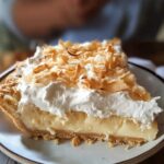 Homemade coconut cream meringue pie slice with golden crust, creamy coconut filling, fluffy meringue, and toasted coconut flakes on top.