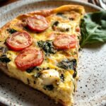 Homemade Frittata Recipe slice with cherry tomatoes, spinach, and cheese on a rustic ceramic plate, garnished with a fresh spinach leaf.
