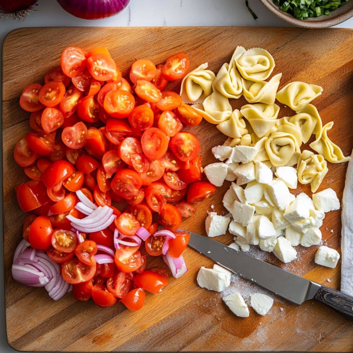 Chopped cherry tomatoes, red onion, mozzarella, and cheese tortellini on a wooden cutting board with a kitchen knife.