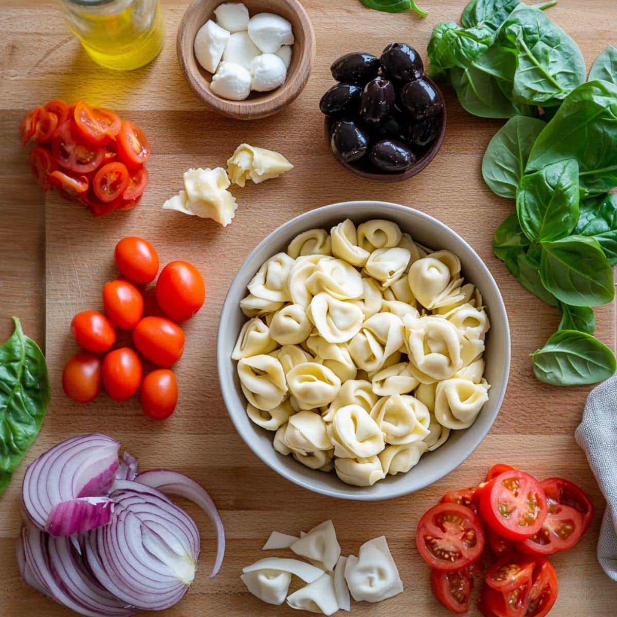 Fresh tortellini pasta salad ingredients on a wooden board, including cheese tortellini, cherry tomatoes, red onion, mozzarella, olives, spinach, and basil.