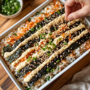 Hand garnishing a sushi bake topped with seaweed strips, spicy mayo, masago, sesame seeds, and green onions in a white casserole dish on a wooden surface.