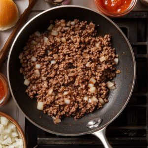 Ground beef and diced onions cooking in a skillet on the stove, surrounded by sauce, raw onions, and a slider bun.