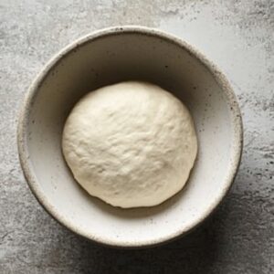 A ball of homemade pizza dough in a ceramic bowl on a gray countertop, ready to rise.