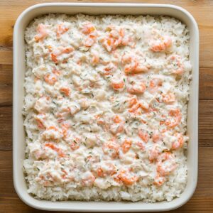 Homemade sushi bake in a white casserole dish with a layer of rice topped with creamy crab and shrimp mixture, ready to bake.