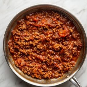 Homemade meat sauce with ground beef, tomatoes, and onions simmering in a stainless steel skillet on a marble countertop.