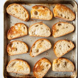Toasted slices of rustic Italian bread on a worn baking sheet, golden and crisp with airy centers.