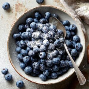 Fresh blueberries in a ceramic bowl lightly coated with flour, with a spoon resting inside and a few berries scattered on a rustic countertop.
