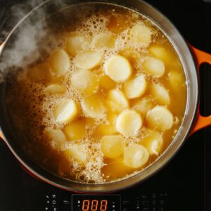 Sliced potatoes simmering in golden chicken broth inside a cream and orange Dutch oven on a stovetop, with steam rising as part of homemade Zuppa Toscana soup prep.