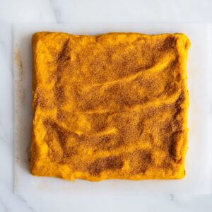 Pumpkin cinnamon roll dough pressed into a square and sprinkled generously with a cinnamon-sugar mixture, ready to be rolled up on a clean white surface.