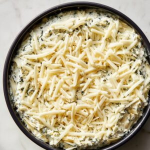 Unbaked spinach artichoke dip in a round black baking dish, topped generously with shredded mozzarella cheese. The creamy dip base with visible spinach is spread evenly beneath the cheese.