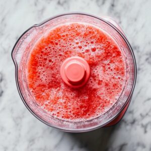 Blended watermelon mixture in a clear food processor with a red lid, foamy and vibrant, on a marble surface.