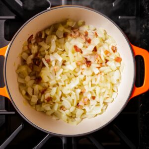 Diced onions sautéing with bits of bacon in a cream-colored Dutch oven with orange handles, set on a black stovetop, gently cooking in fat until soft.