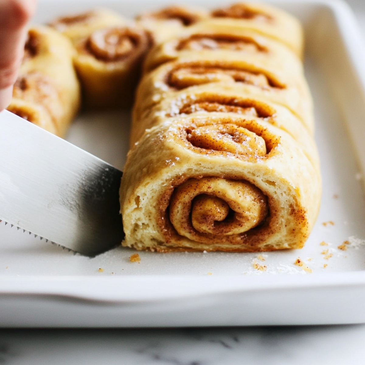 Freshly sliced pumpkin cinnamon rolls arranged in a baking dish, with one roll being cut cleanly by a serrated knife—soft dough swirled with cinnamon-sugar filling in a cozy kitchen scene.