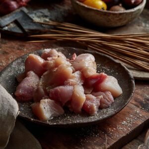 Raw chicken chunks seasoned with salt and pepper on a rustic dark plate, with wooden skewers and natural kitchen tools in the background, ready for homemade ranch garlic parmesan chicken skewers.