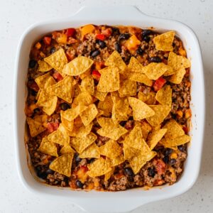 Partially assembled Dorito casserole in a white baking dish with a visible layer of seasoned ground beef, black beans, melted cheese, diced tomatoes, and a topping of whole nacho cheese tortilla chips.
