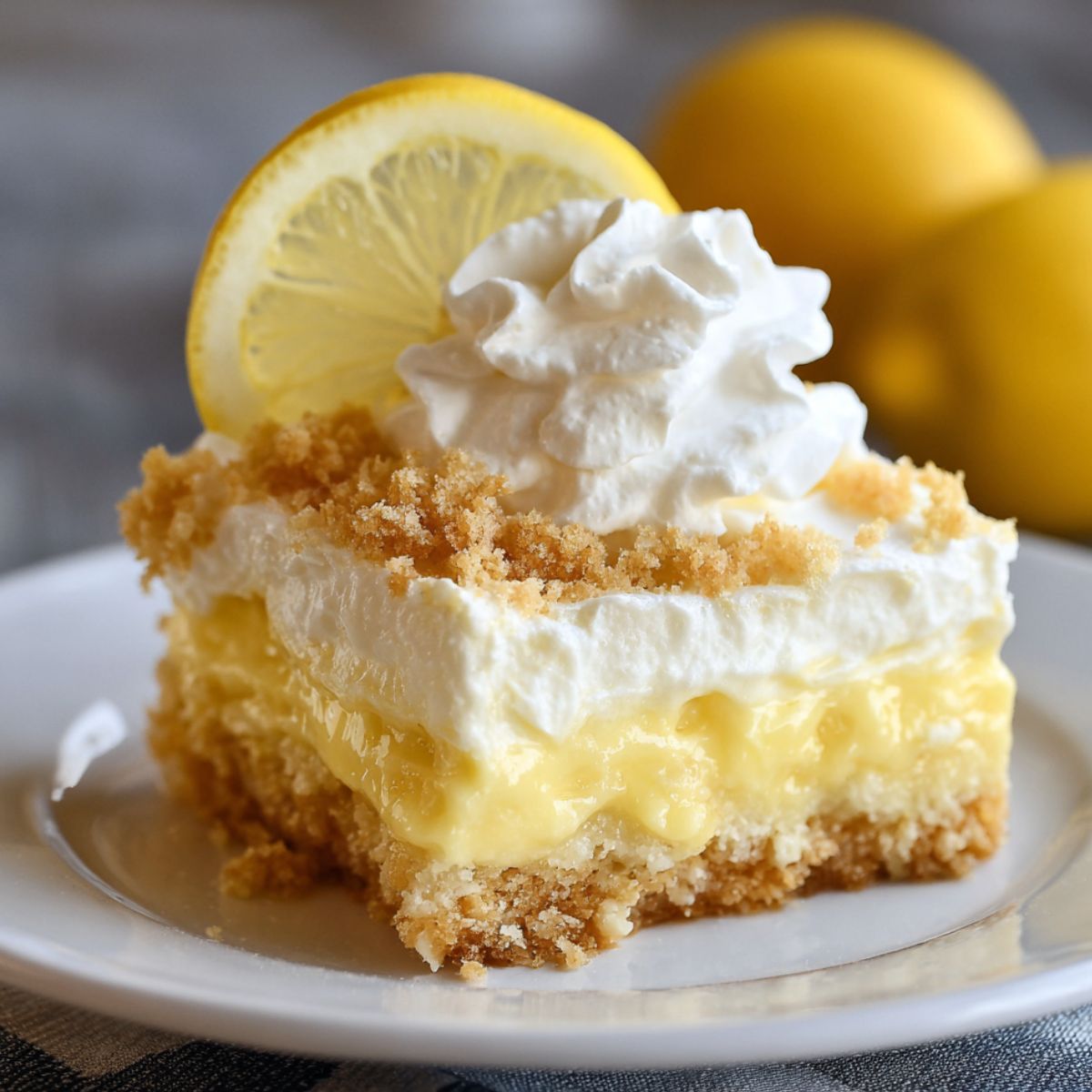 A slice of homemade lemon cream cheese dump cake on a white plate, featuring a crumbly golden crust, creamy lemon filling, whipped topping, and a dollop of whipped cream, garnished with a lemon slice.