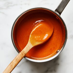 Homemade buffalo sauce in a saucepan being stirred with a wooden spoon, glossy and rich in color, ready for coating crispy cauliflower.