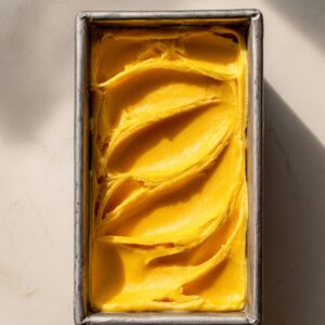 Freshly made mango sorbet in a metal loaf pan, smoothed with soft swirls across the surface. The vibrant yellow-orange sorbet sits on a light marble surface under natural sunlight.
