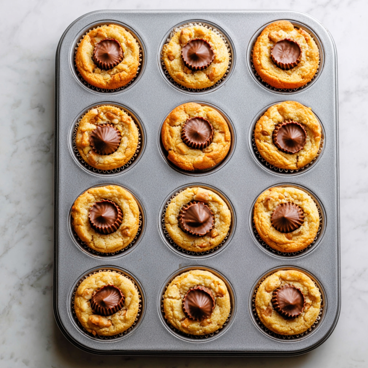 Freshly baked peanut butter cup cookies in a muffin tin, each golden cookie topped with a slightly melted mini peanut butter cup pressed into the center, still warm from the oven.