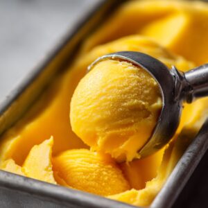 Close-up of a metal ice cream scooper lifting a smooth, round scoop of bright yellow mango sorbet from a rectangular metal container. The texture is creamy with visible frost, and the background is softly blurred.
