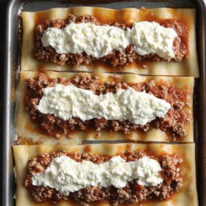 Top-down view of lasagna noodles topped with meat sauce and ricotta, laid flat on a metal tray, ready to roll.
