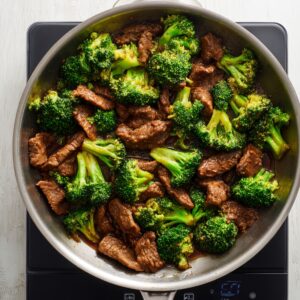 Top-down view of homemade Chinese beef and broccoli in a stainless steel skillet on a black induction cooktop. Tender beef strips in glossy brown sauce are mixed with vibrant green broccoli florets, all glistening under bright natural light.