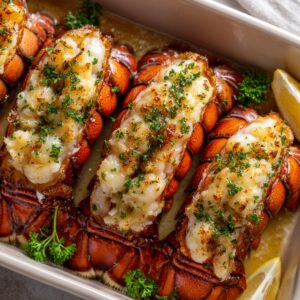 Baked garlic butter lobster tails in a white dish, with golden meat topped with parsley, vibrant red shells, and lemon wedges on the side.