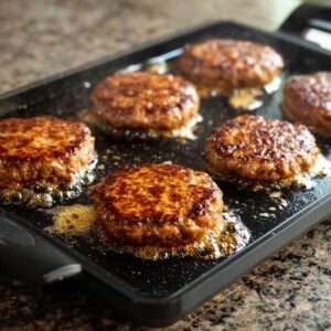 Juicy beef patties sizzling on a black griddle with golden-brown crusts and bubbling grease, set on a granite countertop in a real homemade kitchen scene.