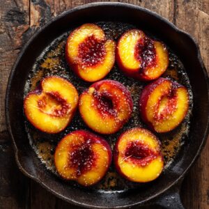 Overhead of peach halves with red centers sizzling in butter in a cast iron skillet on a rustic wooden table.