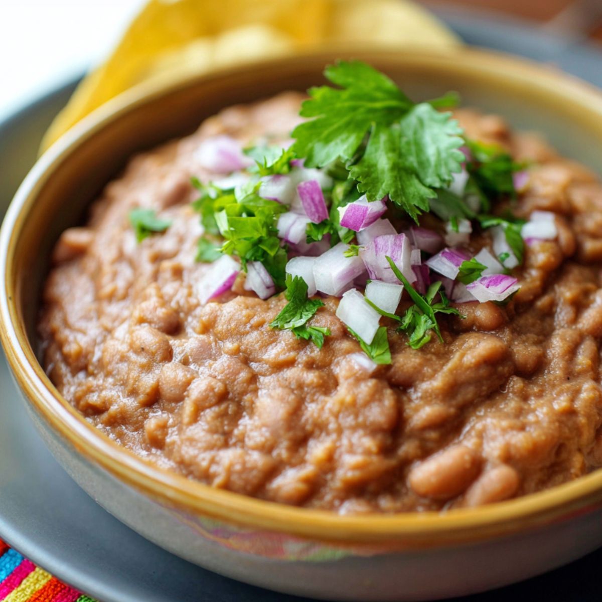 Homemade refried beans served in a rustic ceramic bowl, topped with fresh cilantro and diced red onion, with golden tortilla chips in the background on a gray plate.