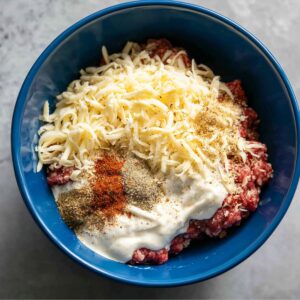 Unmixed ground beef, shredded cheese, mayo, and spices in a blue bowl, ready to be mixed for homemade crack burger patties.
