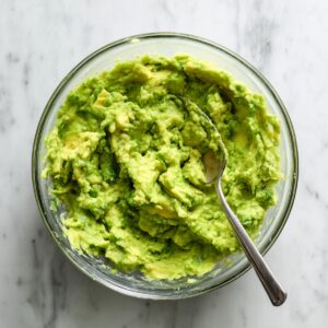 Glass bowl of freshly mashed, chunky avocado with a spoon, on a light gray marble countertop, in natural daylight.