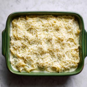 Homemade seafood lasagna in a green ceramic dish, layered with creamy white sauce and pasta, topped with shredded mozzarella and parmesan, sitting on a marble countertop before baking.