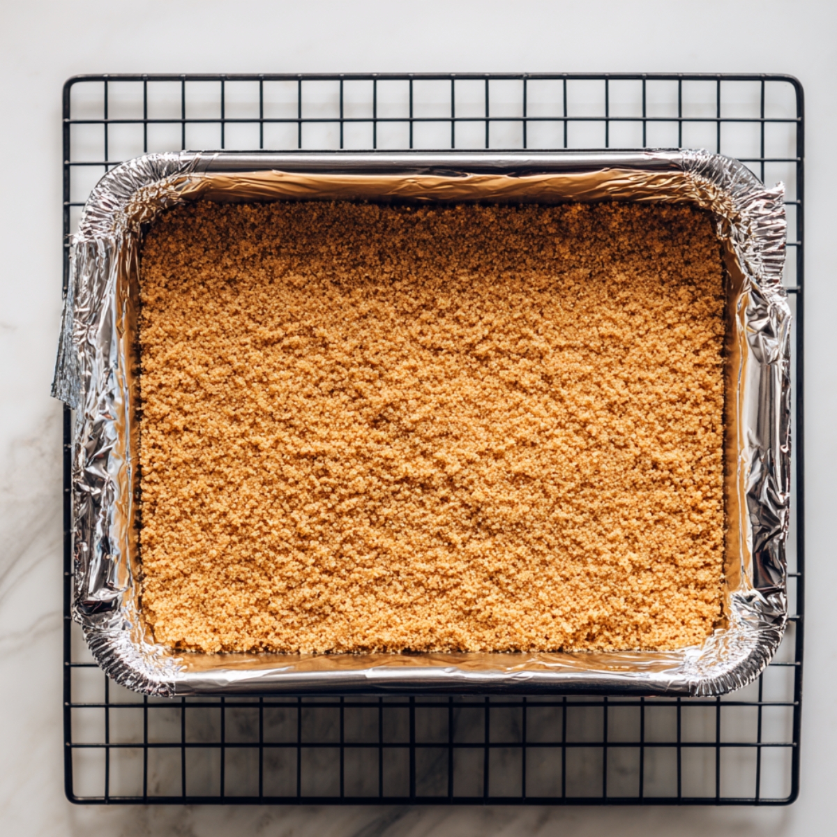 Unbaked graham cracker crust in a foil-lined pan on a cooling rack over a marble surface, smooth and golden, ready for baking.