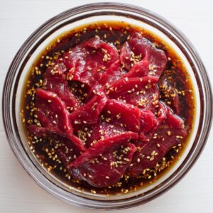 Top-down view of thinly sliced raw beef marinating in a soy-based sauce with sesame seeds in a glass bowl on a white wooden surface, bright natural light.