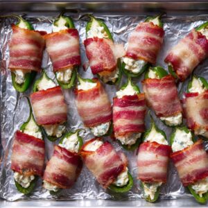 Unbaked jalapeño poppers stuffed with cheese and wrapped in bacon on a foil-lined baking sheet.