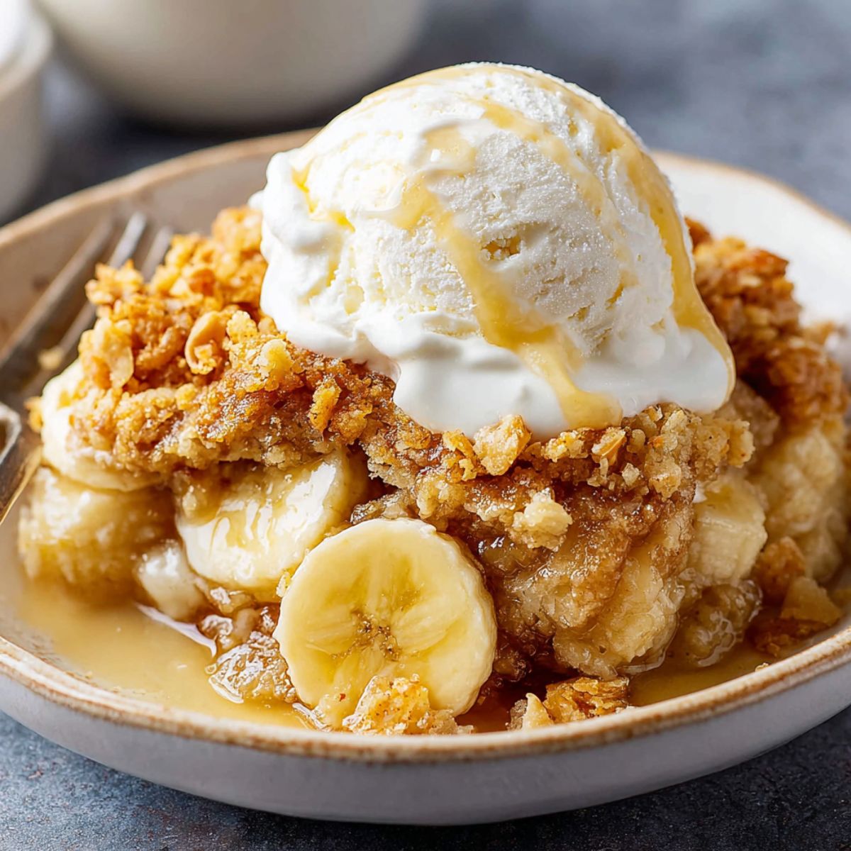 Banana Cobbler Recipe topped with vanilla ice cream and syrup, with visible banana slices and a golden crumb topping. A spoon rests in the bowl, and a whole banana is in the background.
