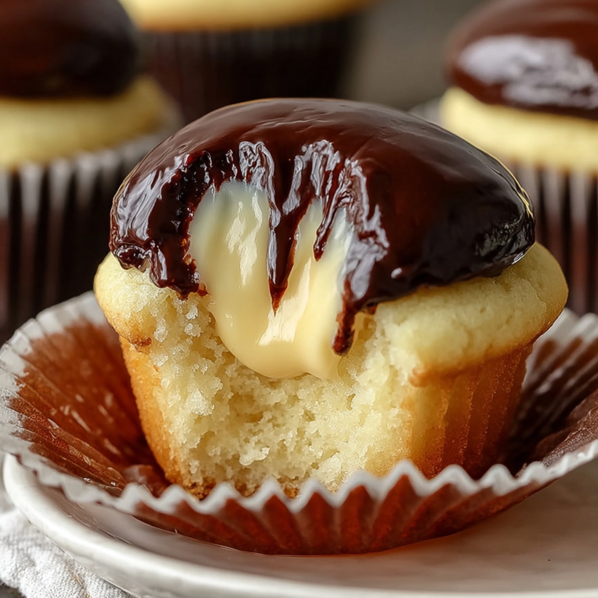 Best Boston Cream Cupcakes Recipe with a bite taken out, showing creamy custard filling dripping from the soft vanilla cake, topped with a thick, glossy layer of chocolate ganache.
