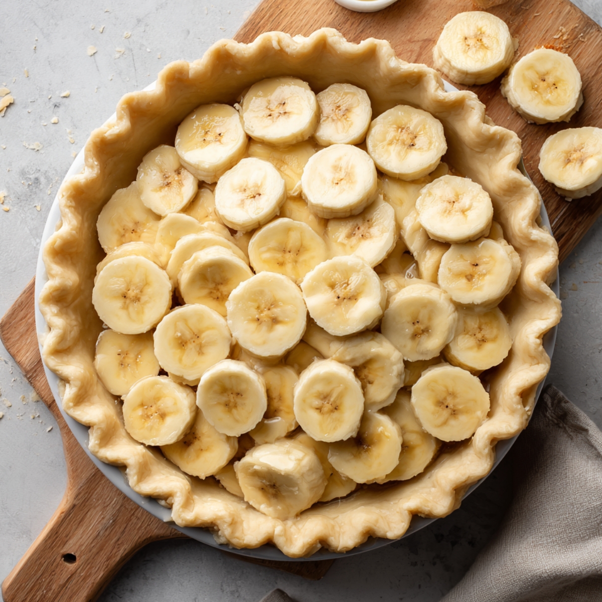 Homemade banana cream pie crust filled with fresh banana slices, on a wooden board over a gray surface with extra slices nearby.