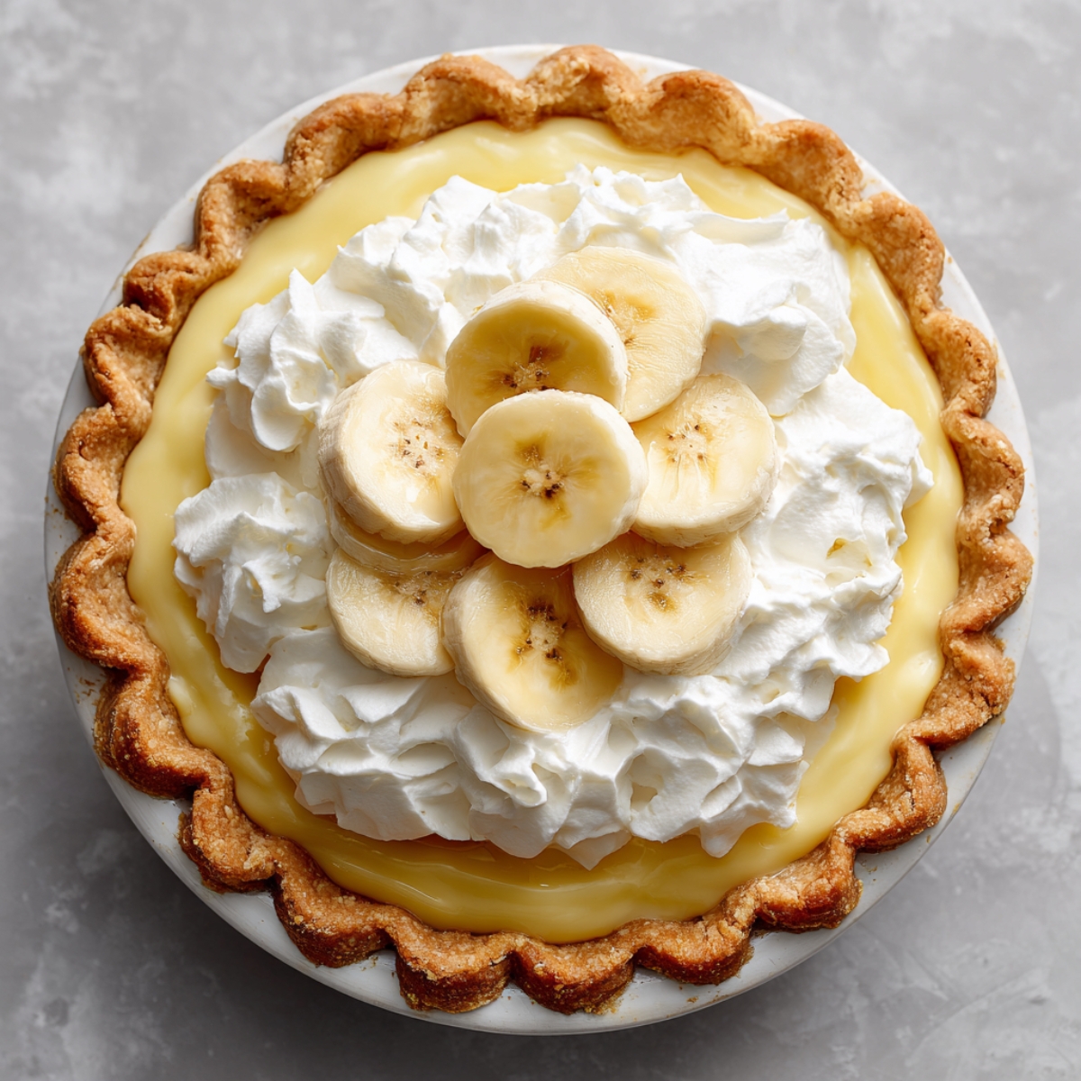 Homemade banana cream pie with golden crimped crust, creamy yellow custard, fluffy whipped cream, and fresh banana slices on top, shot overhead on a gray surface.