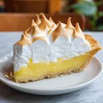 Homemade Lemon Meringue Pie on a white plate with golden crust, bright yellow lemon curd filling, and tall fluffy meringue peaks toasted golden brown.