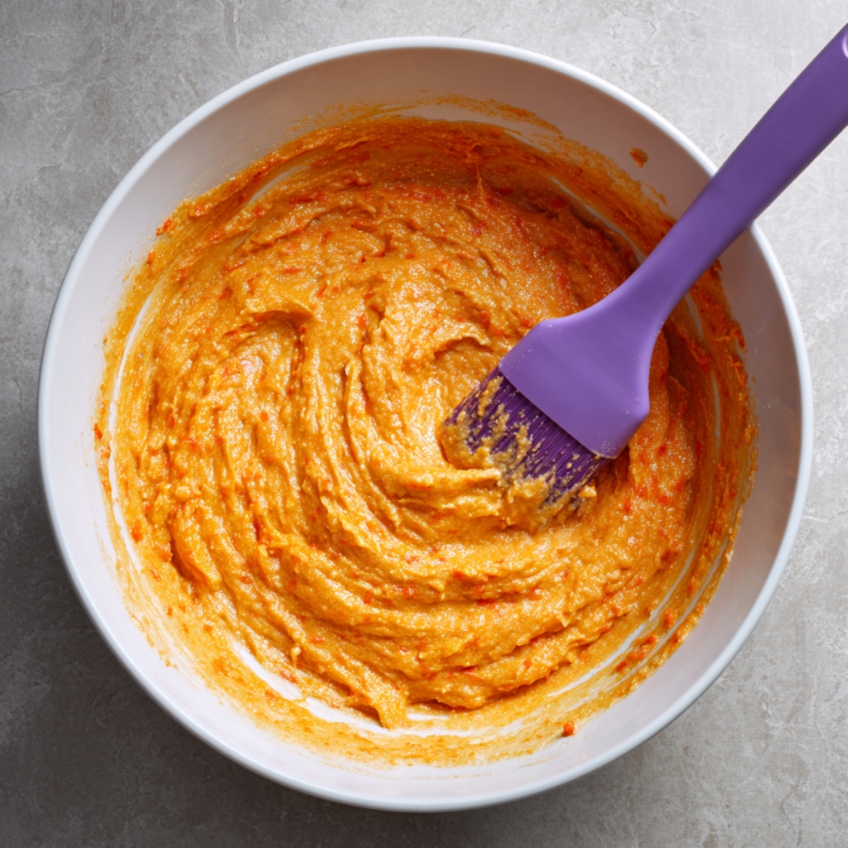 Thick, orange carrot cake batter with visible carrot flecks in a white bowl, mixed with a purple silicone spatula. The bowl sits on a gray stone surface, giving a homemade look.