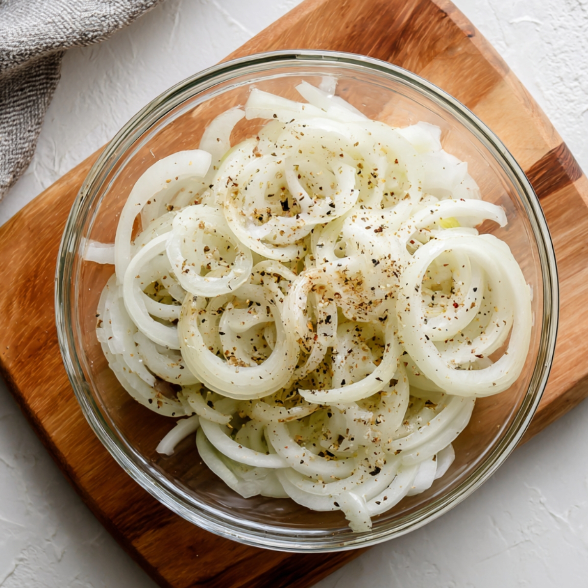 Glass bowl of sliced onions seasoned with salt and pepper on a wooden cutting board.